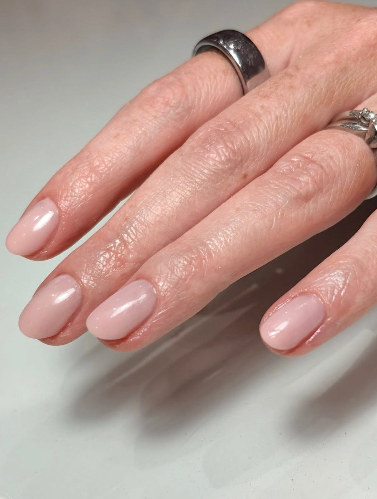 A close-up of a hand with a glossy, light pink manicure, wearing a textured silver band ring on one finger and a silver-toned ring with a clear gemstone on another.