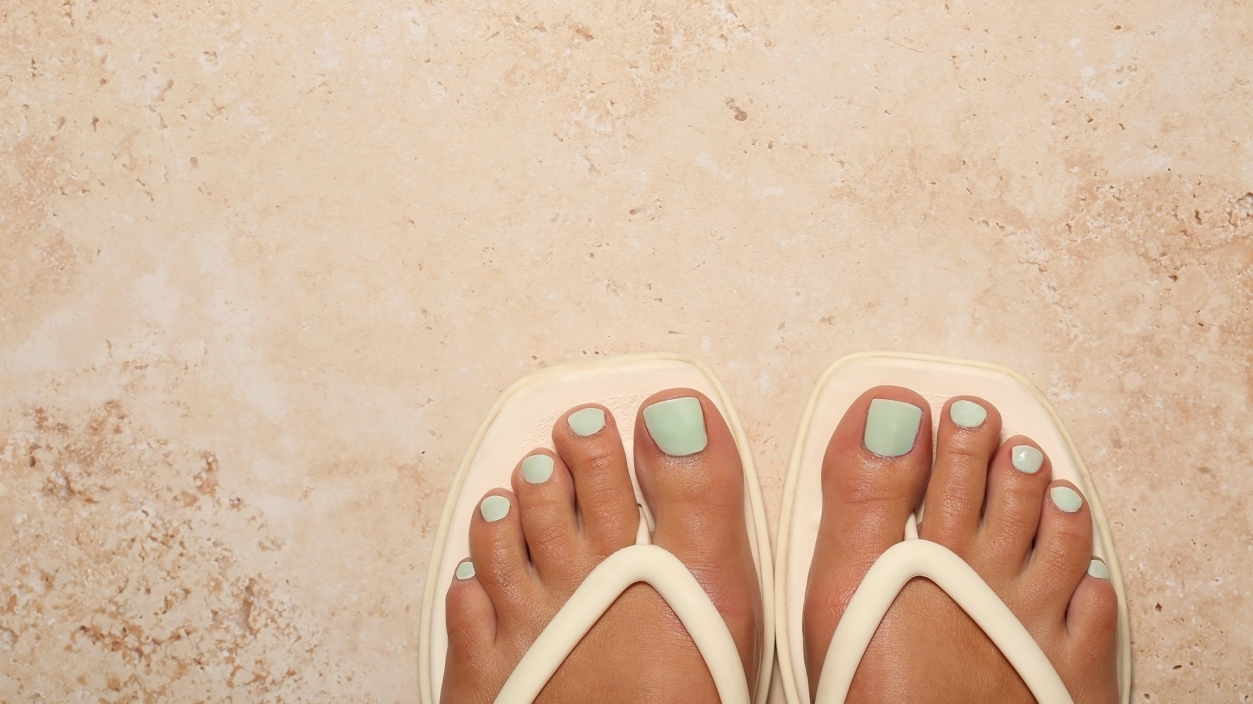 Beige flip-flops worn on feet with light green nail polish on a beige stone floor.