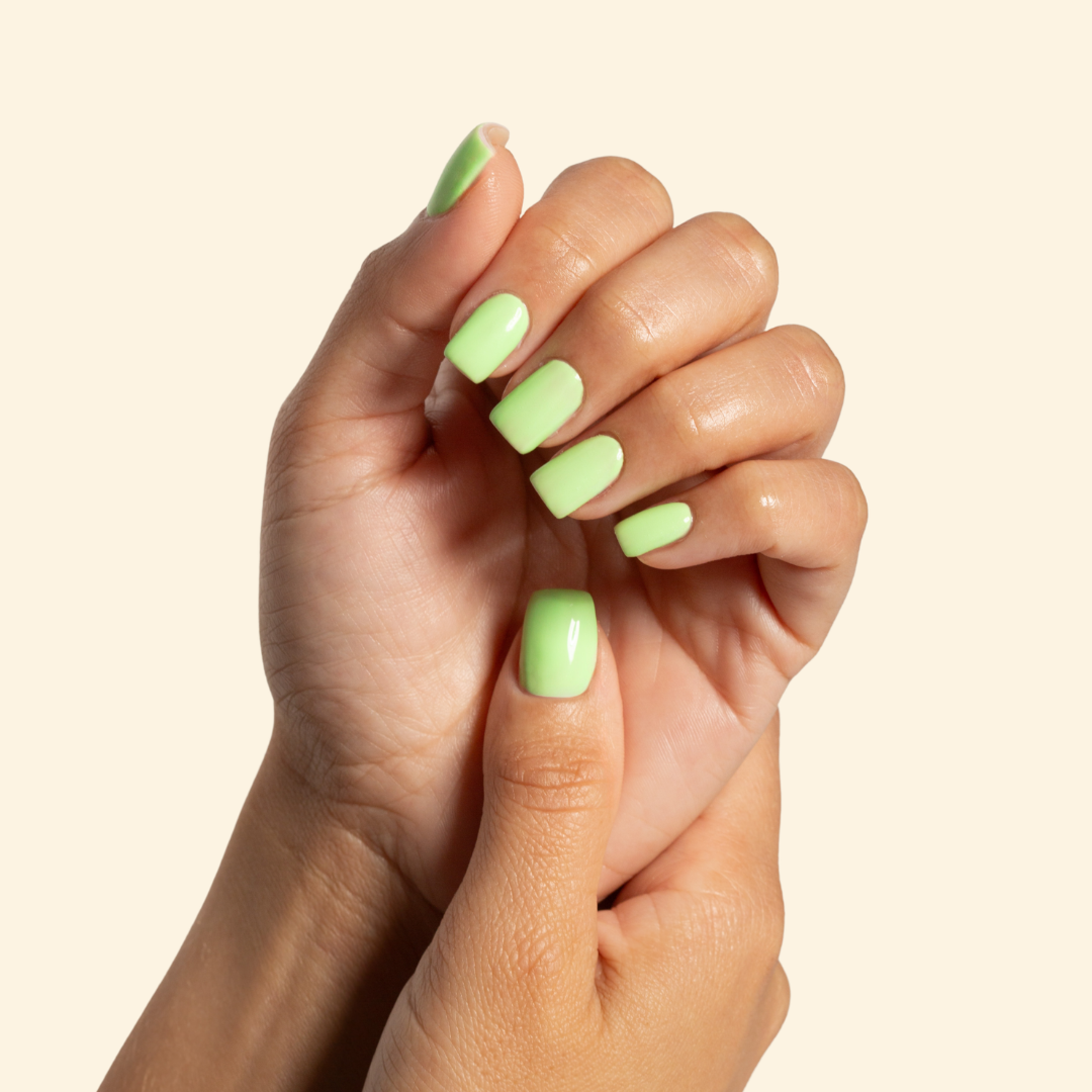 A close-up of two hands with manicured fingernails painted in a glossy light lime green, set against a solid pale cream background.